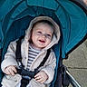 Reginald is registered to the contest to win money with this photo: baby, smiling, stroller, jacket, hood, happy, infant, child, seatbelt, outdoor, pavement, person, cute, blue, white_pants, hands, face, portrait, baby_clothing, daylight