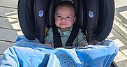 Carson is registered to the contest to win money with this photo: baby, car_seat, blanket, blue_blanket, smile, infant, sunlight, porch, wooden_floor, white_railing, shadows, outdoor, clothing, seatbelt, happy, person, child, cute, cozy, infant_face