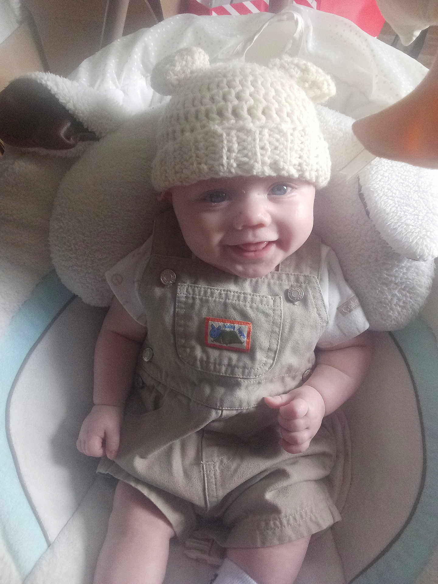 Kaycen is registered to the contest to win money with this photo: baby, smiling, hat, knitted, bear_ears, overalls, beige_clothing, white_shirt, baby_seat, cushion, happy, infant, cute, child, small_hands, indoor, portrait, person, clothing, headwear