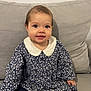 baby, child, girl, sitting, couch, gray_couch, floral_dress, white_collar, socks, polka_dot, indoors, cute, smiling, young_child, person, portrait, happy, comfortable, home, furniture