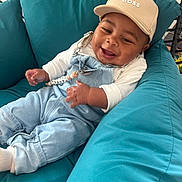 Jayden a rejoint le concours — aidez-le/la à gagner de superbes lots ! baby, child, smile, hat, cap, overalls, blue, white, cushion, chair, indoor, happy, person, infant, clothing, relaxing, cute, portrait, sitting, cozy