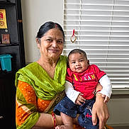 Divit joined the competition — help win amazing prizes! elderly_woman, baby, smiling, indoor, window_blinds, colorful_clothing, holding_baby, portrait, happy, family, love, casual_wear, red_shirt, navy_shorts, bracelets, home, seated, skin_tone, face, person