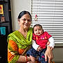 elderly_woman, baby, smiling, indoor, window_blinds, colorful_clothing, holding_baby, portrait, happy, family, love, casual_wear, red_shirt, navy_shorts, bracelets, home, seated, skin_tone, face, person
