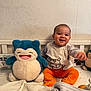 baby, child, plush_toy, smiling, bed, orange_pants, white_top, cartoon_print, happy, indoor, wallpaper, toy, cute, infant, sitting, person, cozy, furniture, soft_toy, room