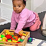 baby, toddler, child, pink_clothing, crawling, toy_fruits, toy_vegetables, wooden_crate, indoor, playtime, curious, colorful, hair_clips, floor_mat, carpet, child_playing, cute, young_child, person, home