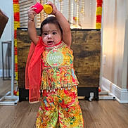 Meera is registered to the contest to win money with this photo: baby, child, paint, colorful, traditional_clothing, floor, wooden_floor, garlands, decorations, indoors, standing, holding, jars, bright_colors, cute, smiling, celebration, festival, portrait, person