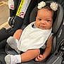 baby, infant, car_seat, bib, socks, curly_hair, flower_headbands, black_car_seat, child, indoor, portrait, cute, smile, small_hands, white_bib, cozy, seated, looking_at_camera, skin, headwear