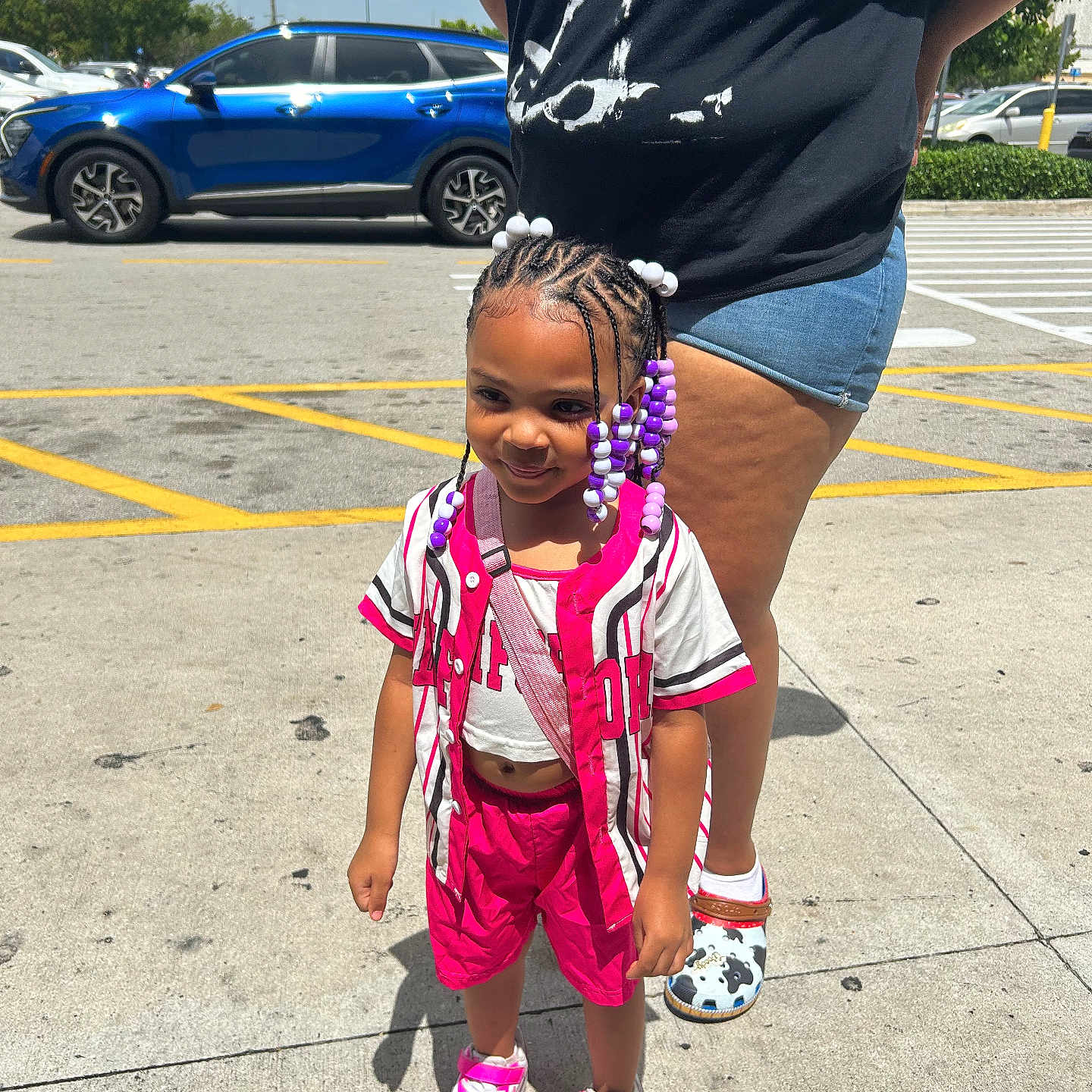 Mikaia is registered to the contest to win money with this photo: adult, beads, braided_hair, car, child, daylight, denim_shorts, footwear, girl, outdoor, parking_lot, person, pink_clothing, shadow, shorts, sidewalk, sneakers, sunny, tshirt, urban