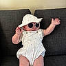 Isla is registered to the contest to win money with this photo: baby, child, sunglasses, hat, white_outfit, couch, indoor, cute, playful, smiling, infant, person, fashion, accessory, seated, furniture, portrait, casual, hands_up, relaxed