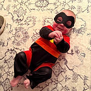 Joey is registered to the contest to win money with this photo: baby, child, superhero_costume, mask, red, black, orange, carpet, pattern, smile, happy, lying_down, feet, hands, indoors, person, costume, cute, playful, infant