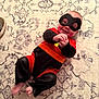 baby, child, superhero_costume, mask, red, black, orange, carpet, pattern, smile, happy, lying_down, feet, hands, indoors, person, costume, cute, playful, infant