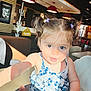baby, blue_dress, chair, child, cute, face, floral_pattern, girl, hair_clips, indoor, person, pigtails, playful, portrait, red_lamps, restaurant, smiling, table, toddler, warm_light