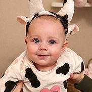 Nina participe au concours pour gagner de l'argent avec cette photo : baby, beige_wall, blue_eyes, cheeks, close_up, cow_costume, cute, french_text, hands_supporting, headband, heart_print, horns, indoor, infant, nursery, onesie, portrait, smile, soft_toys, spot_pattern