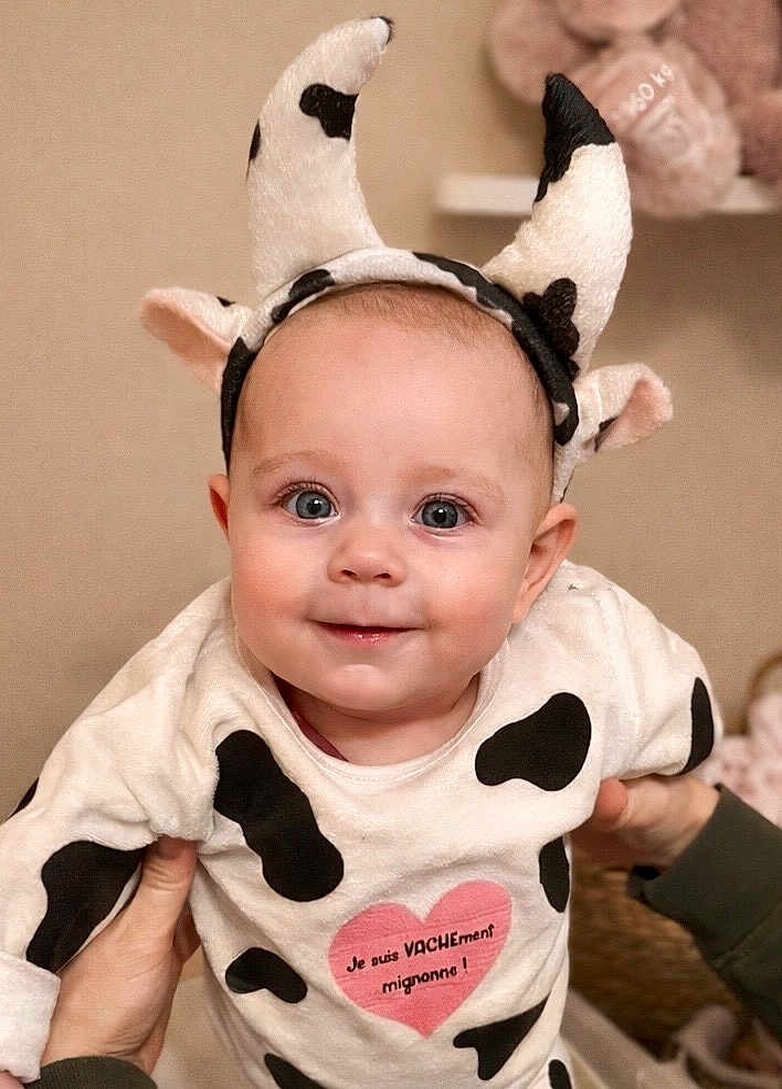 Nina participe au concours pour gagner de l'argent avec cette photo : baby, cow_costume, horns, headband, onesie, smile, blue_eyes, cheeks, portrait, close_up, hands_supporting, indoor, nursery, soft_toys, heart_print, french_text, spot_pattern, beige_wall, cute, infant
