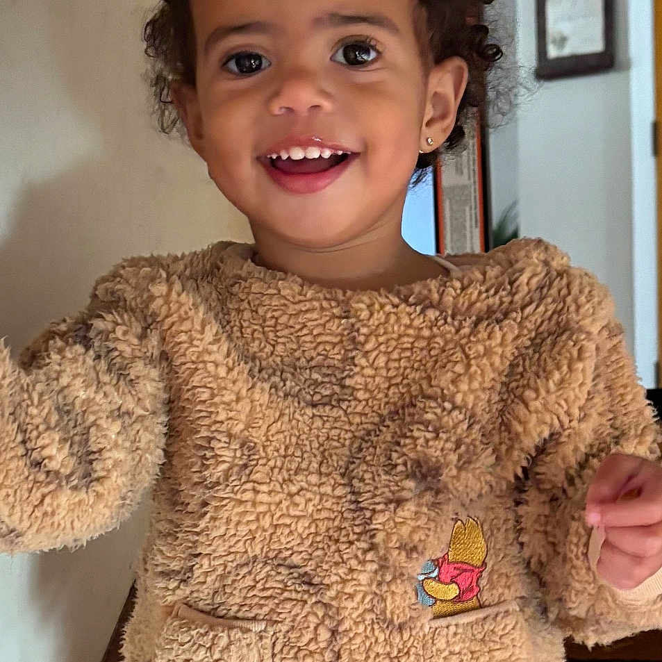 Peyton joined the competition — help win amazing prizes! beige, child, cozy_clothing, curly_hair, cute, door, earrings, embroidered_character, fuzzy_outfit, happy, home, indoor, pigtails, pocket, portrait, smiling, standing, toddler, wall, wood_floor