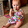 baby, child, toddler, smiling, sitting, indoor, playroom, floral_dress, pink_clothing, teether, toy, bow_shaped_toy, baby_teething, blue_eyes, carpet, books, laundry_basket, furniture, portrait, happy