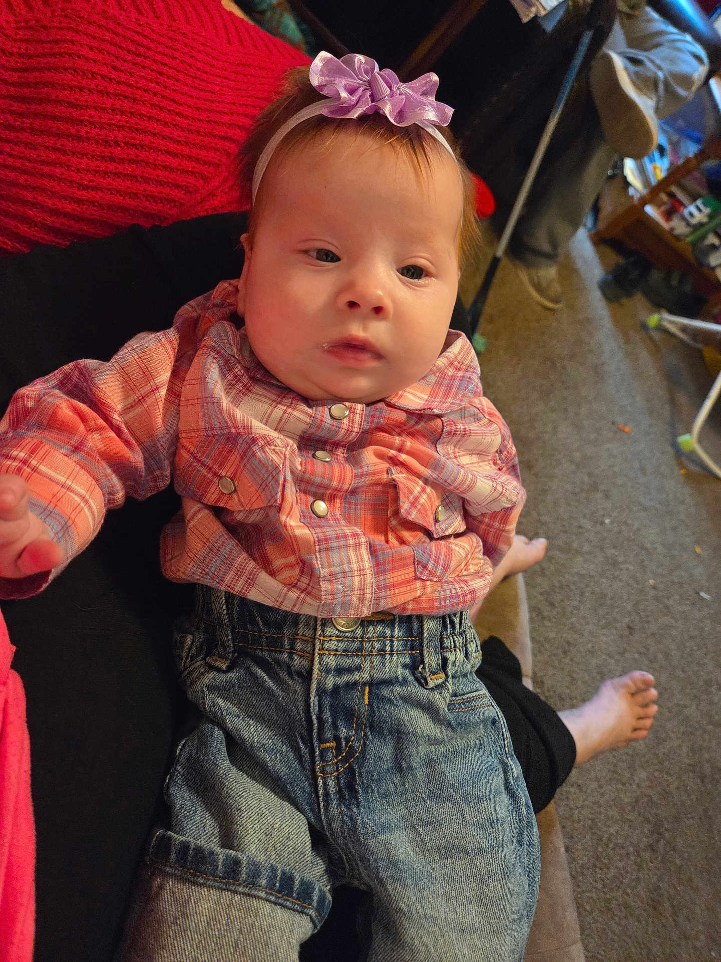 Jade is registered to the contest to win money with this photo: baby, child, plaid_shirt, denim_jeans, headband, purple_bow, person, indoor, carpet, living_room, foot, hand, couch, casual_clothing, seated, infant, expression, young_child, relaxed, soft_lighting
