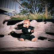 Néva participe au concours pour gagner de l'argent avec cette photo : baby, black_clothing, building, casual, child, curious, cute, daylight, driftwood, face, grass, nature, outdoor, person, sitting, smiling, snack, tree, wood, young