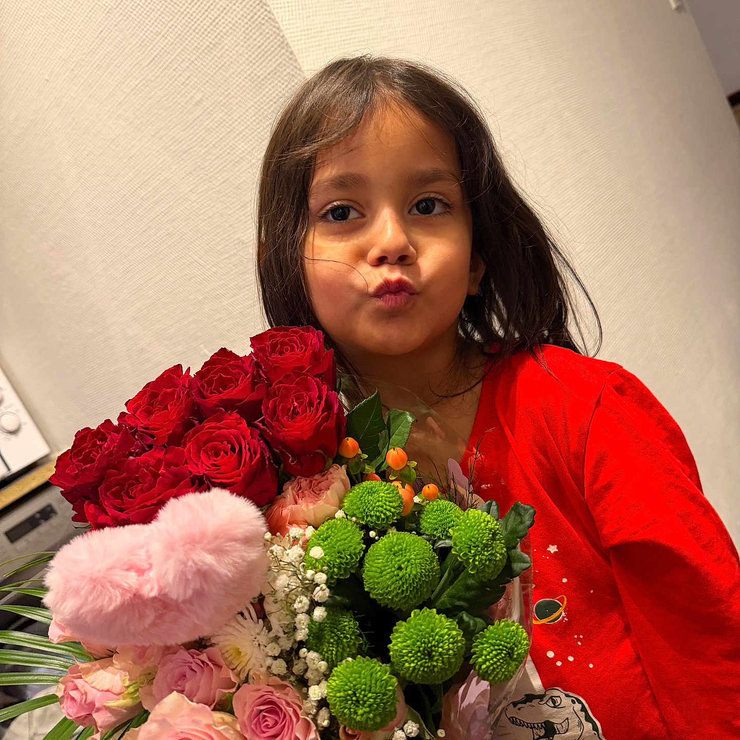 Anais a rejoint le concours — aidez-le/la à gagner de superbes lots ! child, girl, flowers, bouquet, red_shirt, dinosaur_print, puckered_lips, indoor, portrait, dark_hair, green_flowers, pink_roses, red_roses, playful_expression, wall, casual_clothing, person, flower_arrangement, plant, leaf