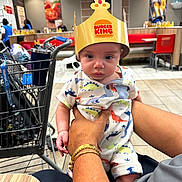 Joni is registered to the contest to win money with this photo: adult_hand, baby, bracelet, burger_king, casual, child, clothing, crown, curious, dinosaur_print, fast_food, floor, food_court, indoor, person, restaurant, seated, shopping_cart, table, tile_floor