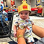 adult_hand, baby, bracelet, burger_king, casual, child, clothing, crown, curious, dinosaur_print, fast_food, floor, food_court, indoor, person, restaurant, seated, shopping_cart, table, tile_floor