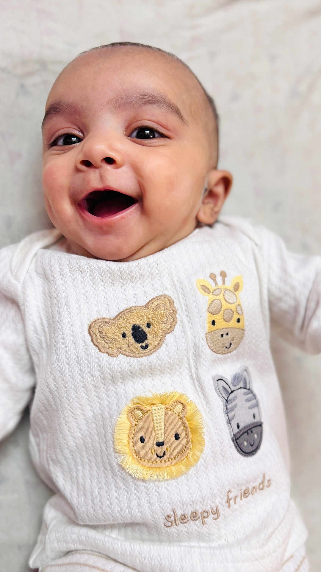 Hunaid is registered to the contest to win money with this photo: baby, smiling, infant, closeup, white_clothing, embroidered_animals, koala, giraffe, lion, zebra, soft_texture, happy, cute, portrait, child, lying_down, indoors, young_child, sleepy_friends, cozy