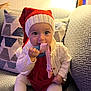baby, child, infant, red_hat, knit_hat, teething_ring, white_sweater, red_dress, tights, couch, pillow, indoor, smiling, cute, holiday, festive, chewing, seated, cozy, warm_lighting