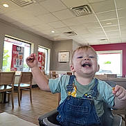 Waylon is registered to the contest to win money with this photo: toddler, child, high_chair, indoor, restaurant, fast_food, smile, happy, messy, food, clothing, denim_overalls, table, chair, window, daylight, ceiling_lights, floor, person, casual