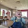 toddler, child, high_chair, indoor, restaurant, fast_food, smile, happy, messy, food, clothing, denim_overalls, table, chair, window, daylight, ceiling_lights, floor, person, casual