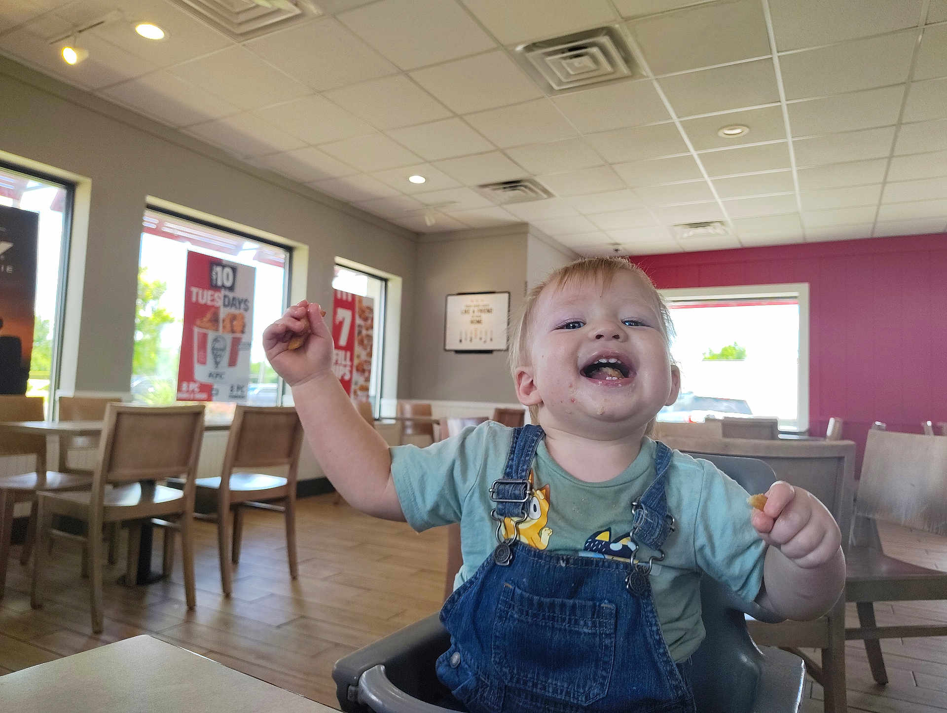 Waylon is registered to the contest to win money with this photo: toddler, child, high_chair, indoor, restaurant, fast_food, smile, happy, messy, food, clothing, denim_overalls, table, chair, window, daylight, ceiling_lights, floor, person, casual