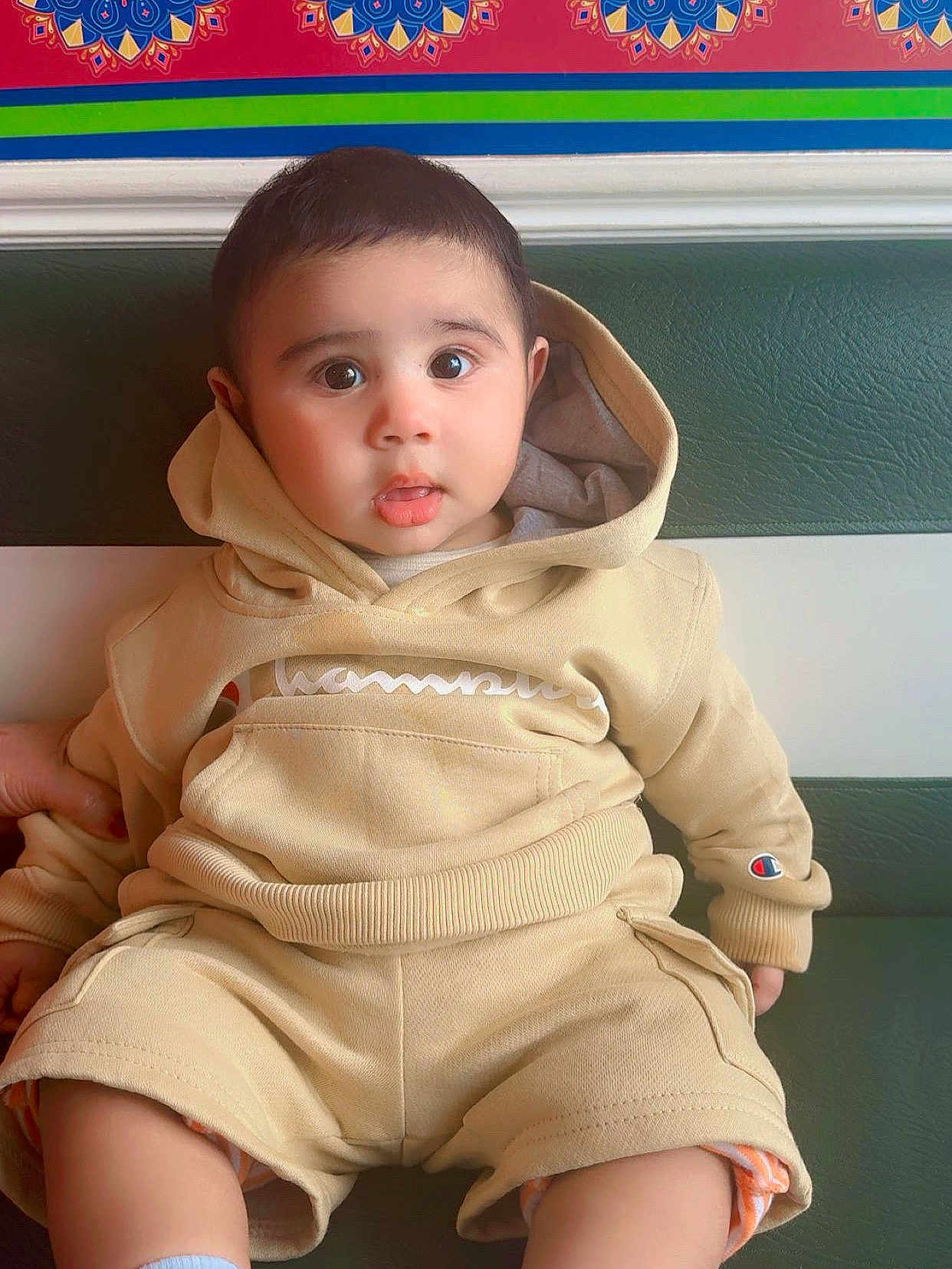 Azan joined the competition — help win amazing prizes! baby, child, hoodie, shorts, sitting, indoors, bench, green, curious, cute, portrait, face, person, clothing, casual, young, expression, comfortable, patterned_wall, colorful