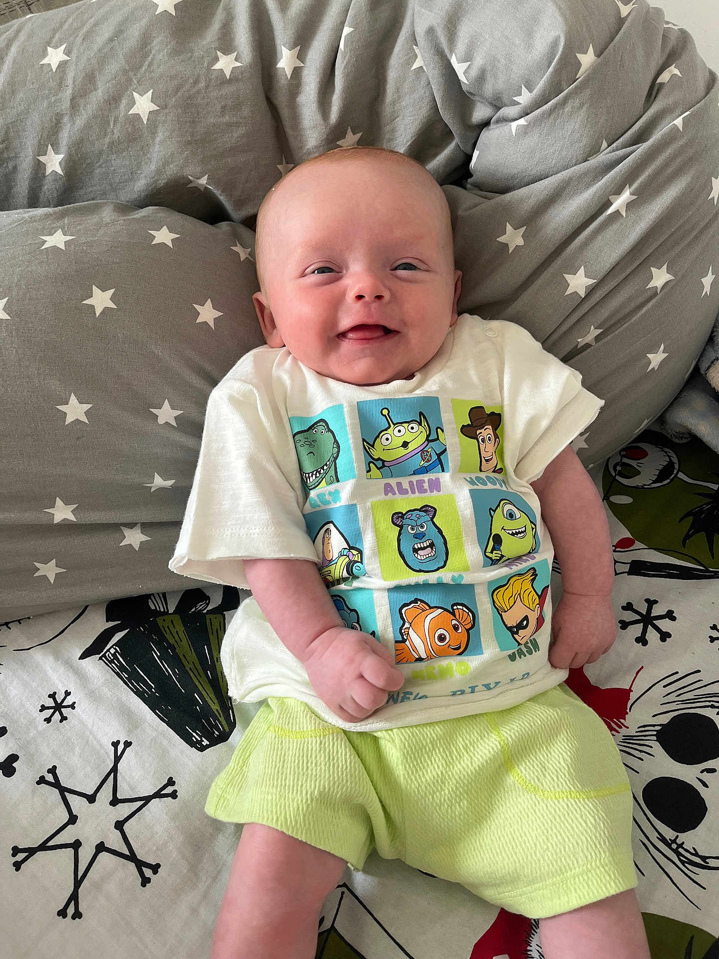 Reggie is registered to the contest to win money with this photo: baby, smiling, tshirt, toy_story, shorts, pillow, stars, bed, blanket, infant, child, cute, happy, indoor, portrait, person, clothing, sitting, face, young