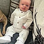 baby, infant, white_clothing, sitting, plush_surface, cushion, smiling, tongue_out, dog_paw, footwear, white_shoes, knitwear, cute, indoors, portrait, child, person, soft_fabric, home, relaxed