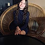 child, girl, smile, long_hair, wavy_hair, sitting, chair, wicker, hands, portrait, indoor, fashion, black_dress, young, person, casual, relaxed, happy, furniture, home