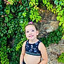 bracelet, casual_clothing, child, daylight, flower, girl, greenery, hair_accessory, happy, leaves, looking_up, nature, outdoor, plant, portrait, rock_wall, sleeveless_top, smile, standing, young