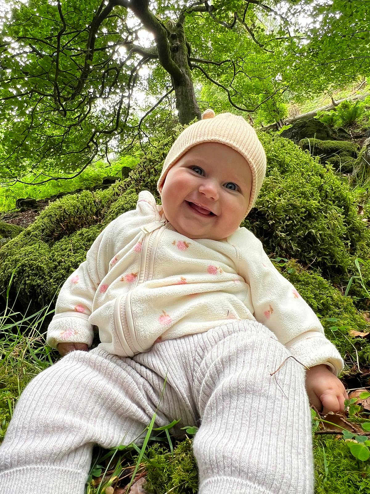 Ellise is registered to the contest to win money with this photo: baby, smiling, forest, moss, greenery, hat, outdoor, nature, child, trees, grass, leaves, happy, cute, infant, clothing, sitting, person, daylight, scenery