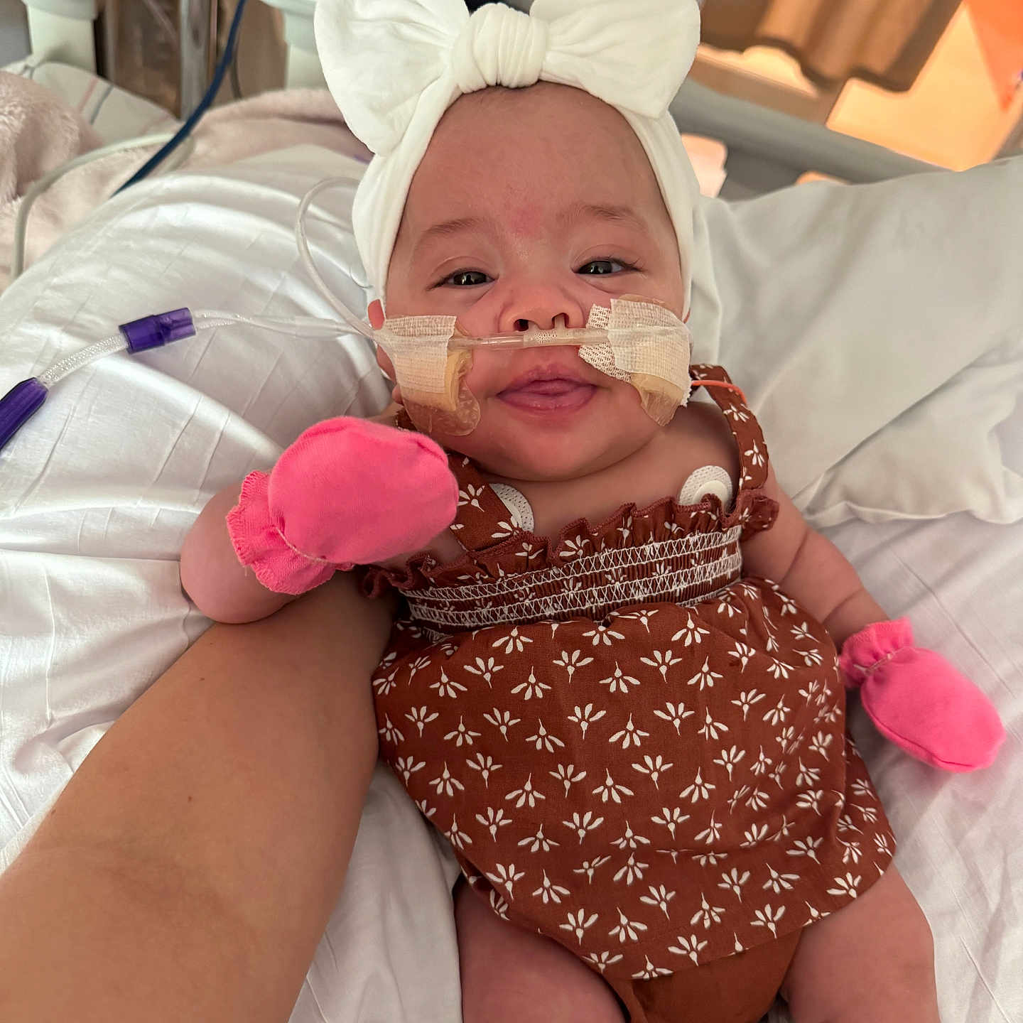 Esmari is registered to the contest to win money with this photo: arm, baby, bed, bow, brown_dress, child, cute, headband, healthcare, hospital, indoors, infant, medical_tubes, mittens, patient, person, smile, support, taped_tubes, white_bedding