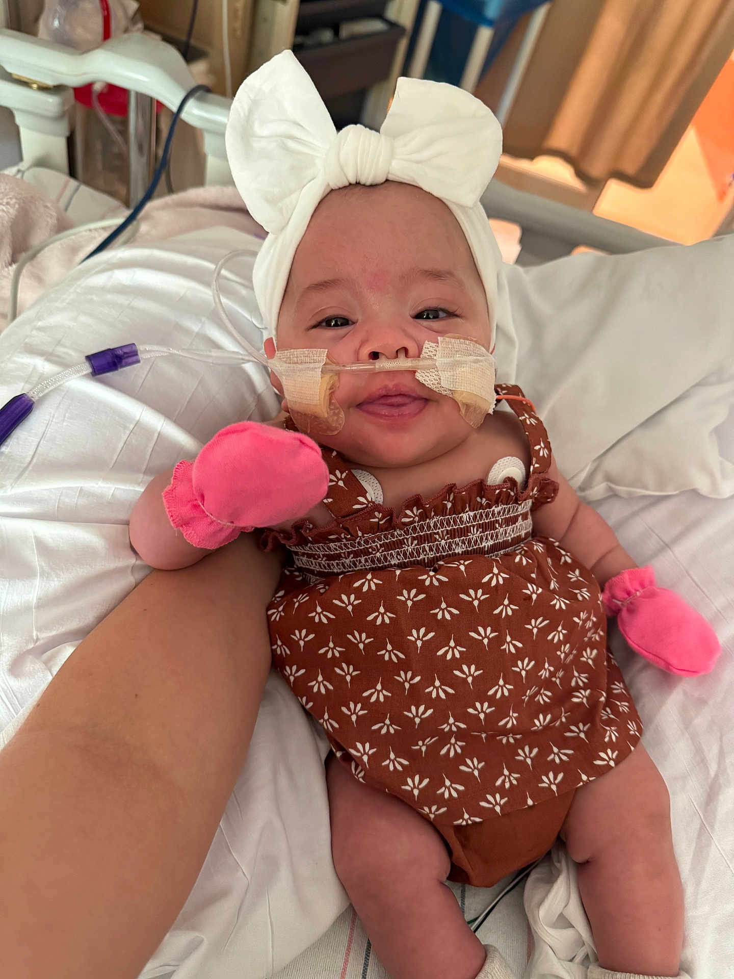 Esmari is registered to the contest to win money with this photo: arm, baby, bed, bow, brown_dress, child, cute, headband, healthcare, hospital, indoors, infant, medical_tubes, mittens, patient, person, smile, support, taped_tubes, white_bedding