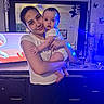 adult, baby, blue_lighting, butterfly_decor, child, clothing, family, furniture, holding, home, indoor, infant, living_room, mother, parent, portrait, short_sleeves, smiling, television, wall_decor