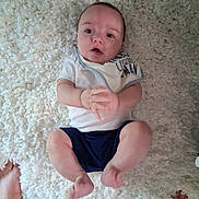 Aryez is registered to the contest to win money with this photo: baby, infant, child, portrait, lying_down, rug, carpet, cream_colored, white_clothing, blue_shorts, hands, feet, wide_eyed, open_mouth, indoors, home, overhead_view, casual_photo, family, safe