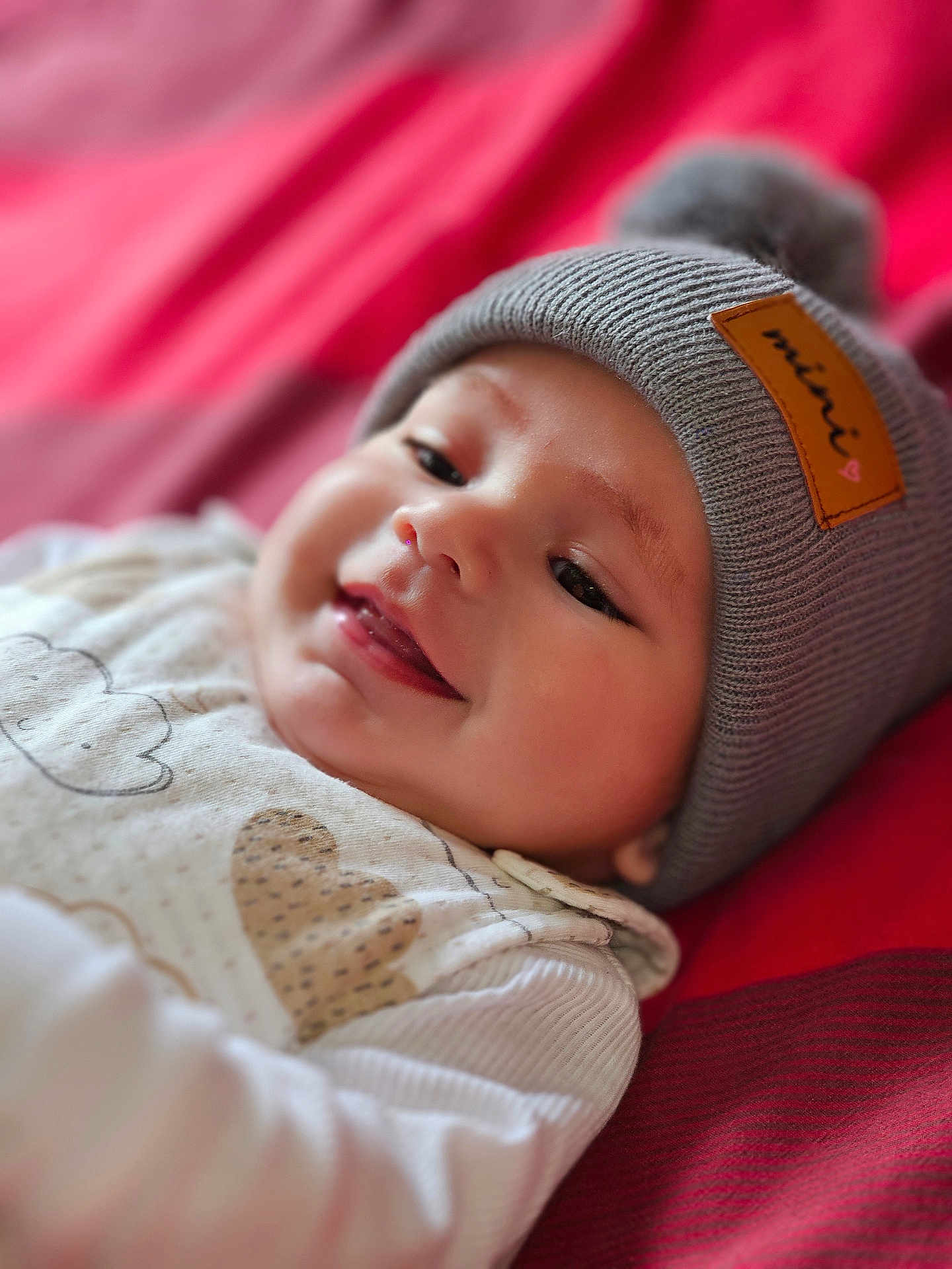 Kenaël participe au concours pour gagner de l'argent avec cette photo : baby, smiling, hat, knit_hat, pom_pom, infant, red_blanket, clothing, close_up, face, happy, cute, portrait, child, lying_down, warm, soft_focus, cozy, indoor, apparel