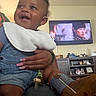 adult_hand, baby, bib, child, denim, furniture, happy, held, home, indoors, infant, living_room, overalls, pacifier, person, portrait, shoes, sitting, smiling, television