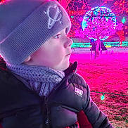 Emilio is registered to the contest to win money with this photo: child, hat, jacket, scarf, winter_clothing, outdoor, night, holiday_lights, tree, illumination, decoration, colorful, festive, person, profile, cold_weather, park, grass, puffer_jacket, curious