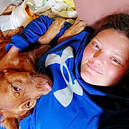 Maple is registered to the contest to win money with this photo: person, dog, blue_sweatshirt, face, lying_down, indoor, pillow, blanket, cozy, pet, cuddling, casual, relaxed, animal, companion, portrait, home, comfort, snuggling, resting