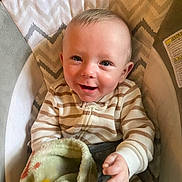 Haden is registered to the contest to win money with this photo: baby, baby_seat, blanket, child, comfort, cozy, cute, eyes, face, furniture, happy, head, indoor, infant, portrait, reaching_hand, smiling, soft_texture, striped_clothing, young_child