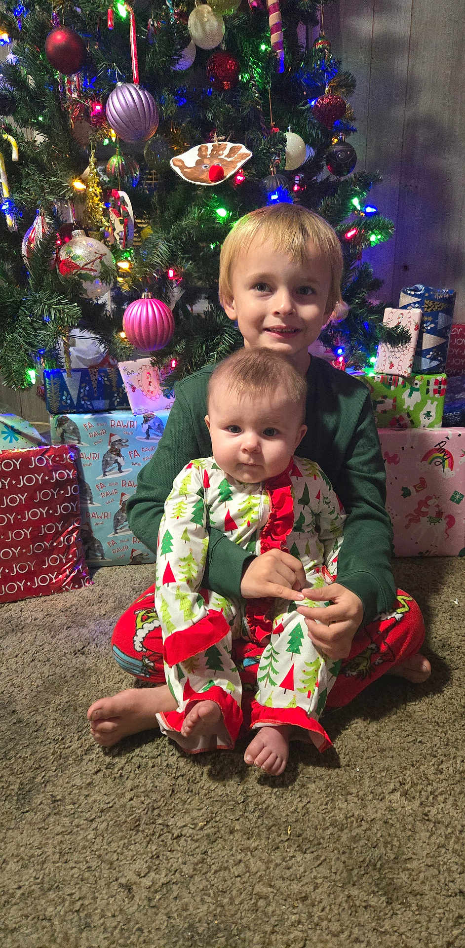 Amy is registered to the contest to win money with this photo: child, baby, sibling, christmas_tree, christmas_ornaments, christmas_lights, presents, pajamas, holiday_pajamas, carpet, indoors, portrait, smiling, festive, wrapped_gifts, ornament, sitting, holiday, young_child, family_moment