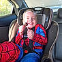 child, spiderman_costume, car_seat, lollipop, smile, sunlight, car_interior, seatbelt, happy, young_child, red, blue, window, seat, fun, face, boy, daylight, candy, safety