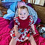 baby, child, smiling, dress, floral, red, blue, blanket, pillow, plush_toy, indoor, cute, seated, happy, face, person, infant, cozy, wood_paneling, curtains
