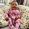 baby, child, smiling, pajamas, hello_kitty, pink, floral_pattern, cushion, indoor, clothing, face, happy, person, portrait, furniture, bedroom, seated, cute, infant, home