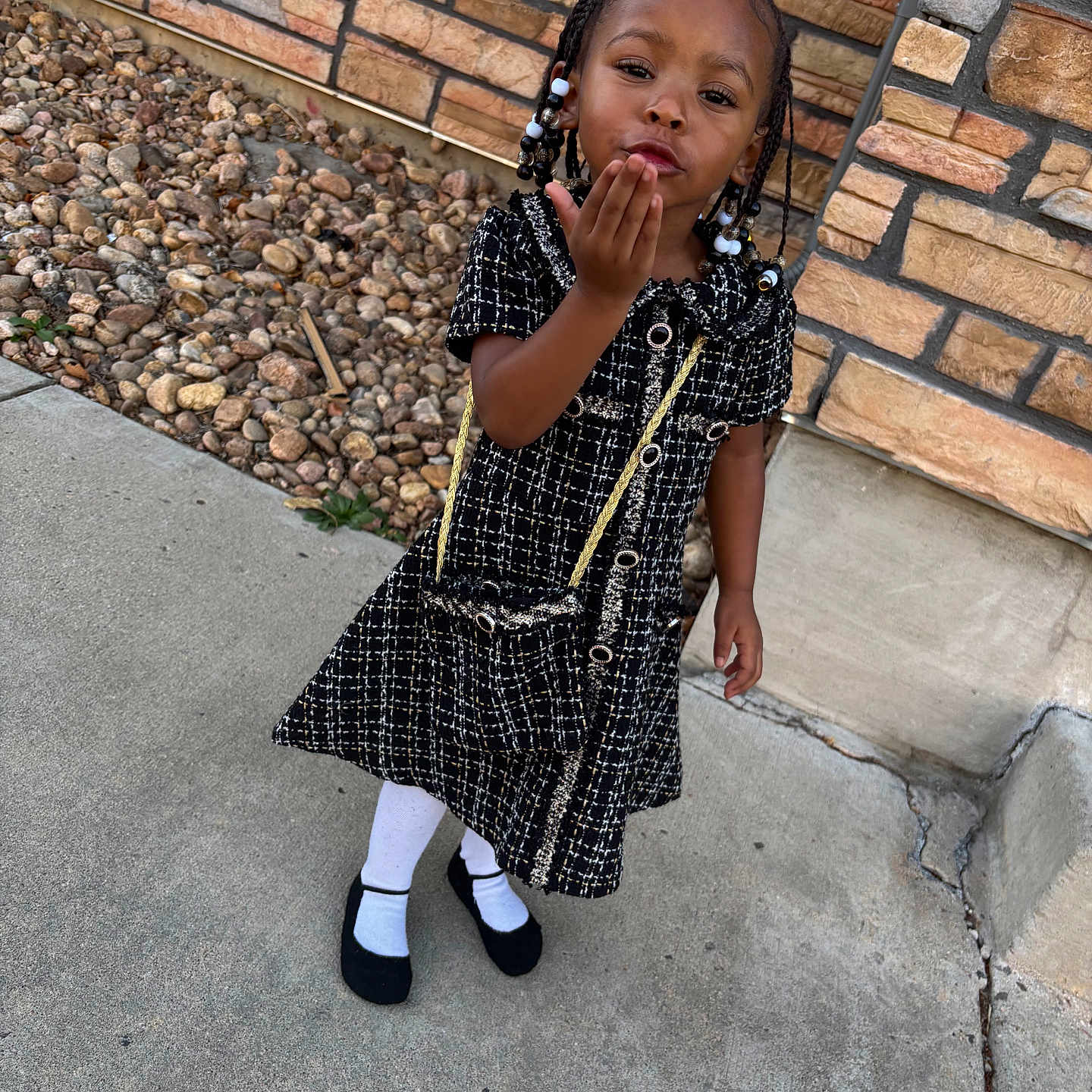 Zyani is registered to the contest to win money with this photo: child, girl, outdoor, dress, black_dress, white_tights, black_shoes, purse, braids, beads, sidewalk, stone_wall, rocks, posing, smiling, cute, fashion, person, young, portrait